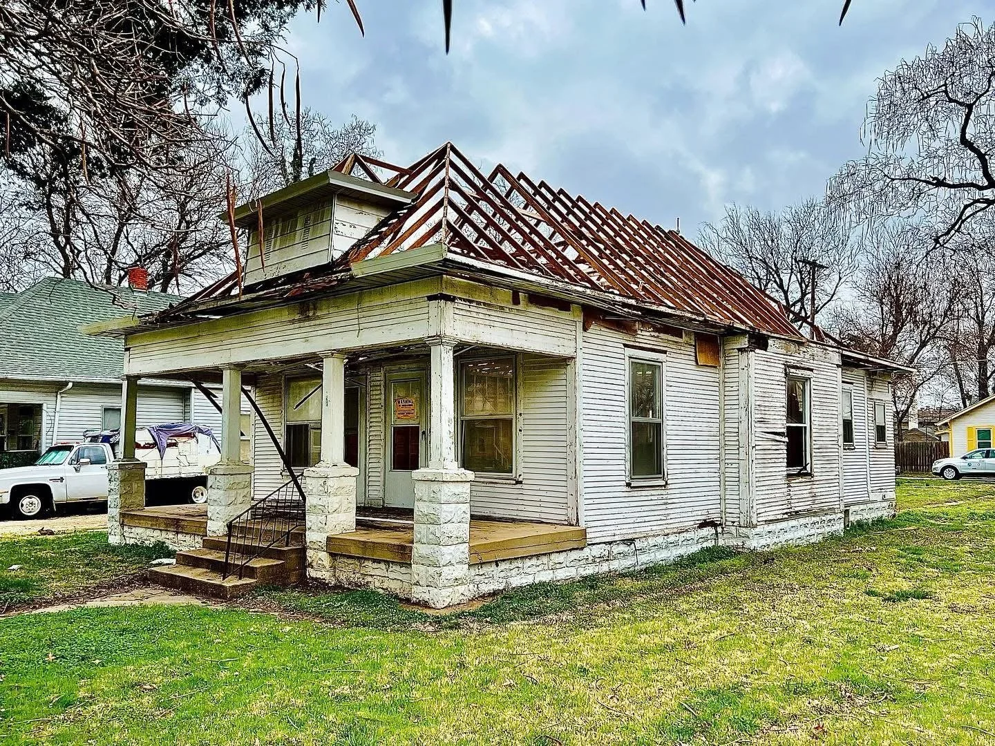 Old, abandoned house with a damaged roof, peeling white paint, and broken windows; front porch with steps and railings, surrounded by grass and leafless trees.