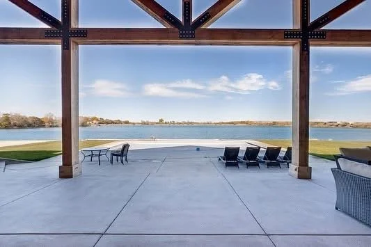 View of a lake or river seen through large open outdoor pavilion with wooden beams, concrete floor, and outdoor furniture including chairs and loungers.