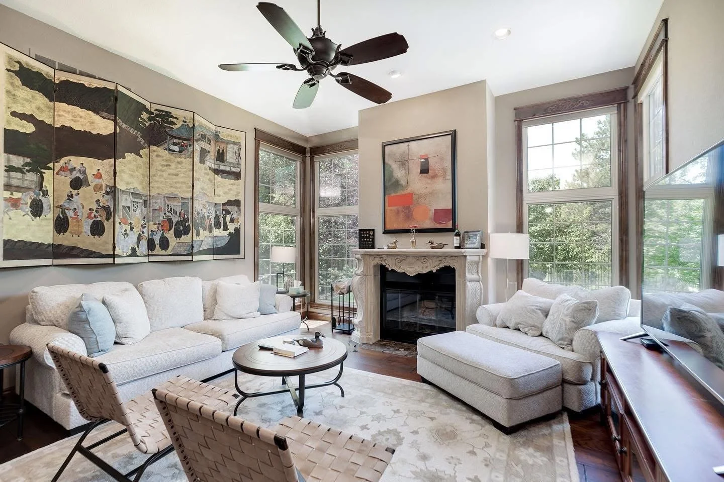 A bright living room with white sofas, a fireplace, large windows, a ceiling fan, and artwork on the walls.