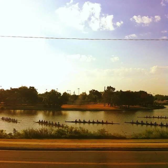 People rowing in single-file boats on a calm river, with trees and a partly cloudy sky in the background, and a road in the foreground.