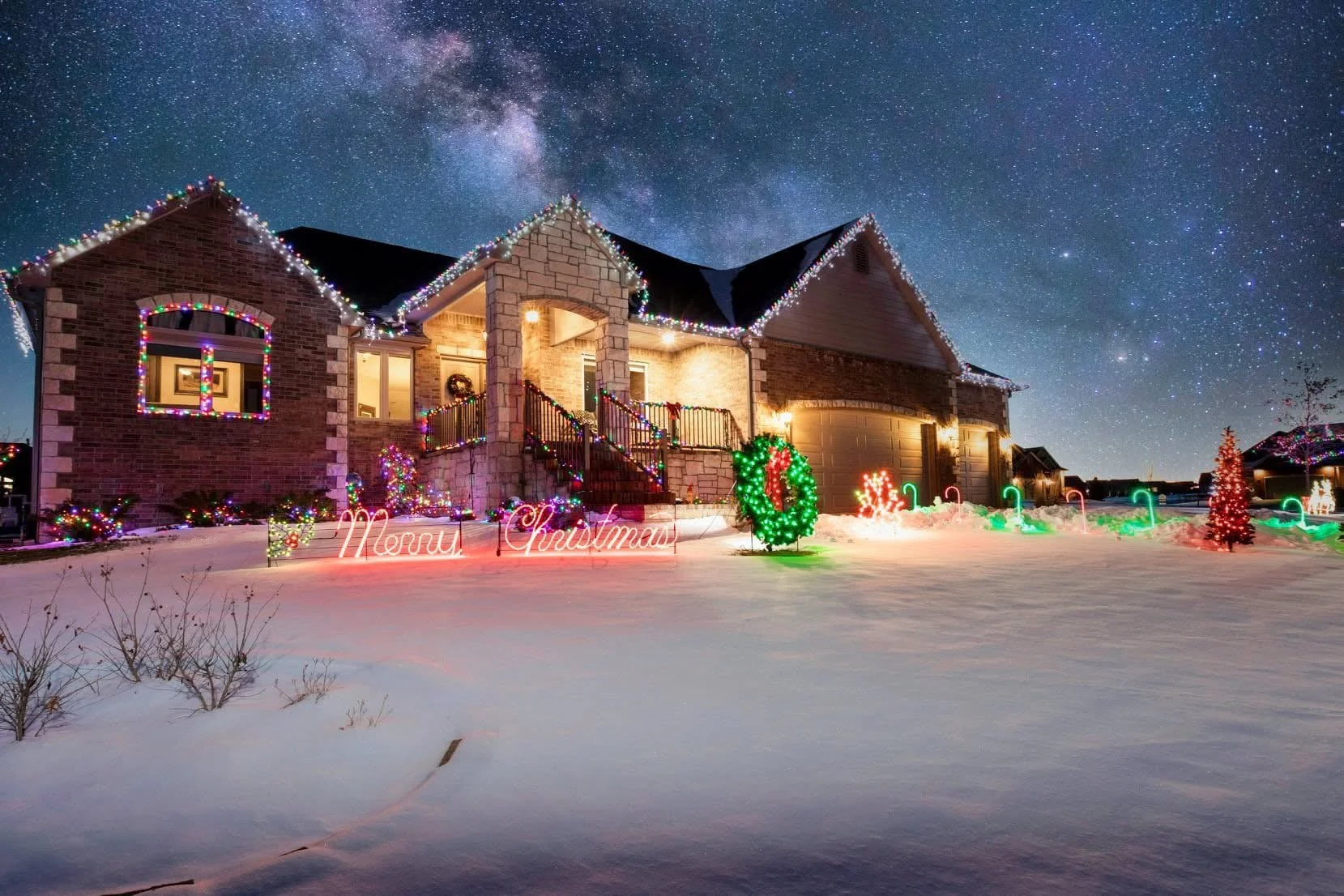 A two-story brick house decorated with colorful Christmas lights under a starry night sky with the Milky Way. Snow covers the ground, and festive holiday signs and decorations, including a wreath and small Christmas trees, are visible outside.