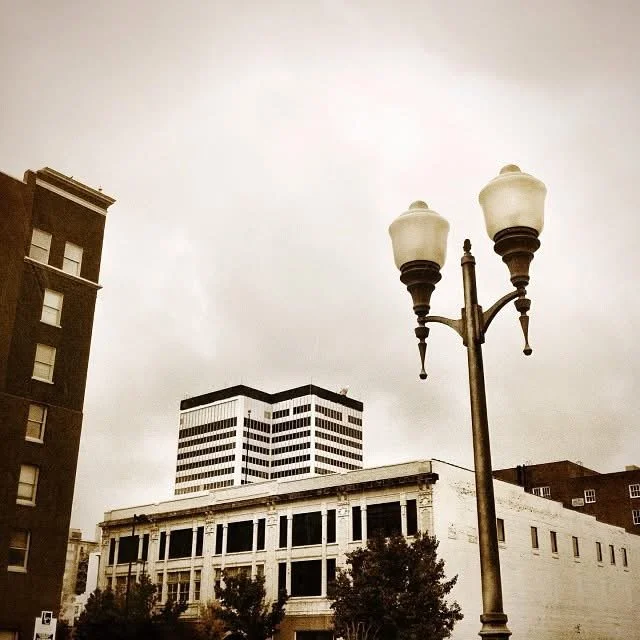 Street scene with a vintage-style street lamp in the foreground, surrounded by older and modern buildings under a cloudy sky.