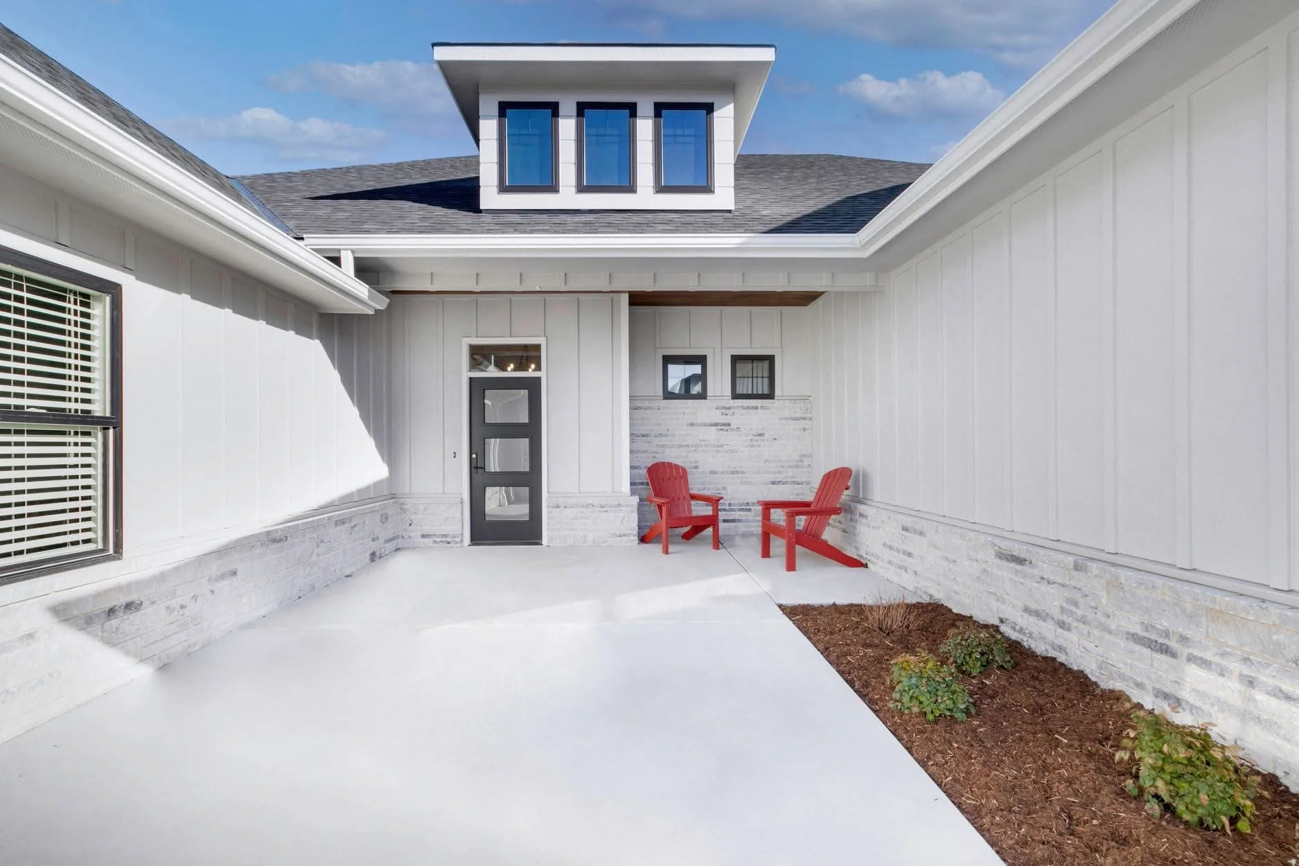 Modern house exterior with a small patio area featuring two red Adirondack chairs, a concrete walkway, white siding, small plants, and a black door with four glass panels.
