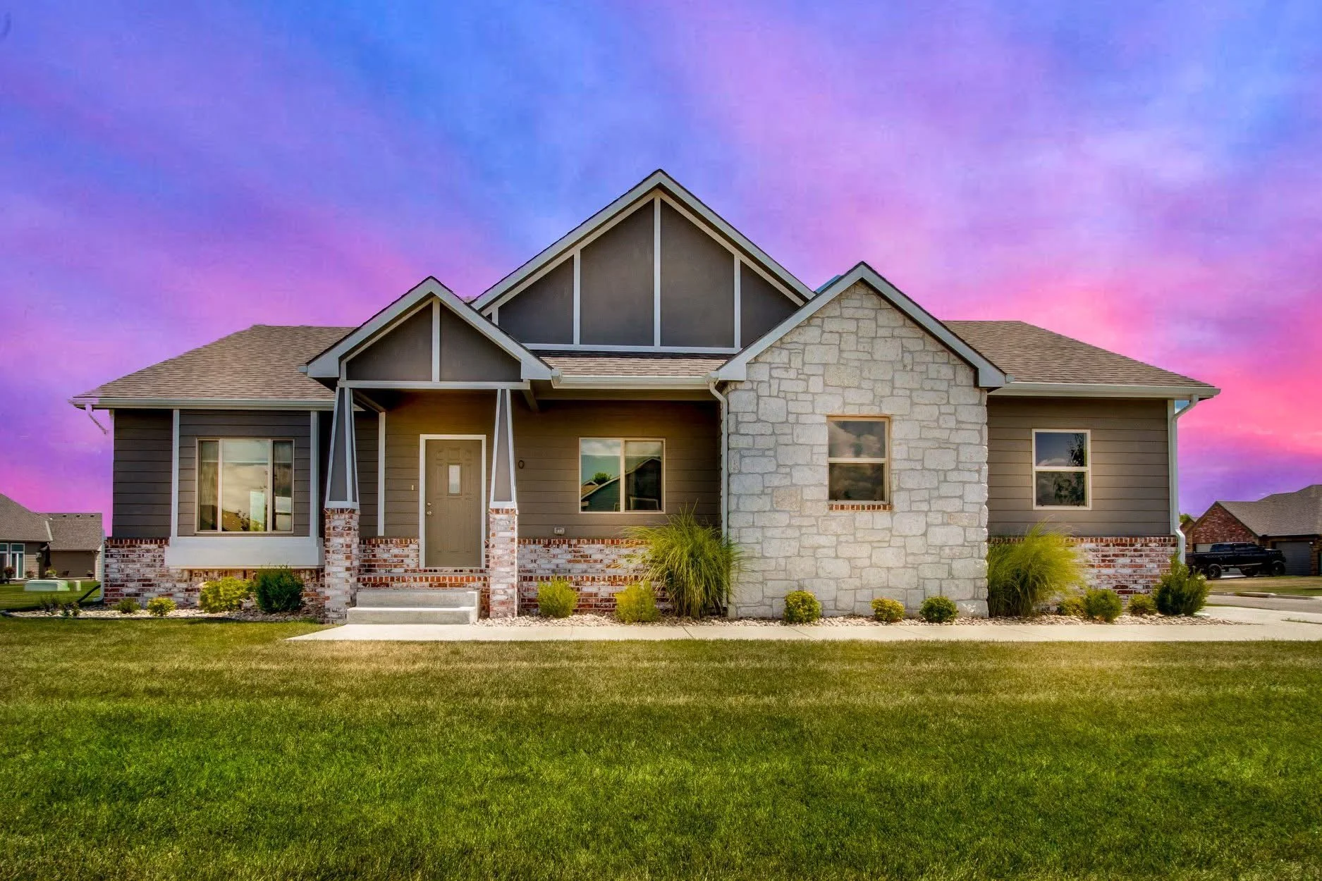 Modern house with gray siding, stone accents, and front porch, set against a vibrant sunset sky, with a well-maintained lawn and garden in the foreground.