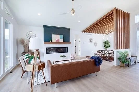 Modern living room with hardwood floors, a brown couch, white walls, and a wall-mounted fireplace. There's a dining area with a wooden table and chairs in the background, a decorative wooden ceiling feature, and large windows providing natural light.