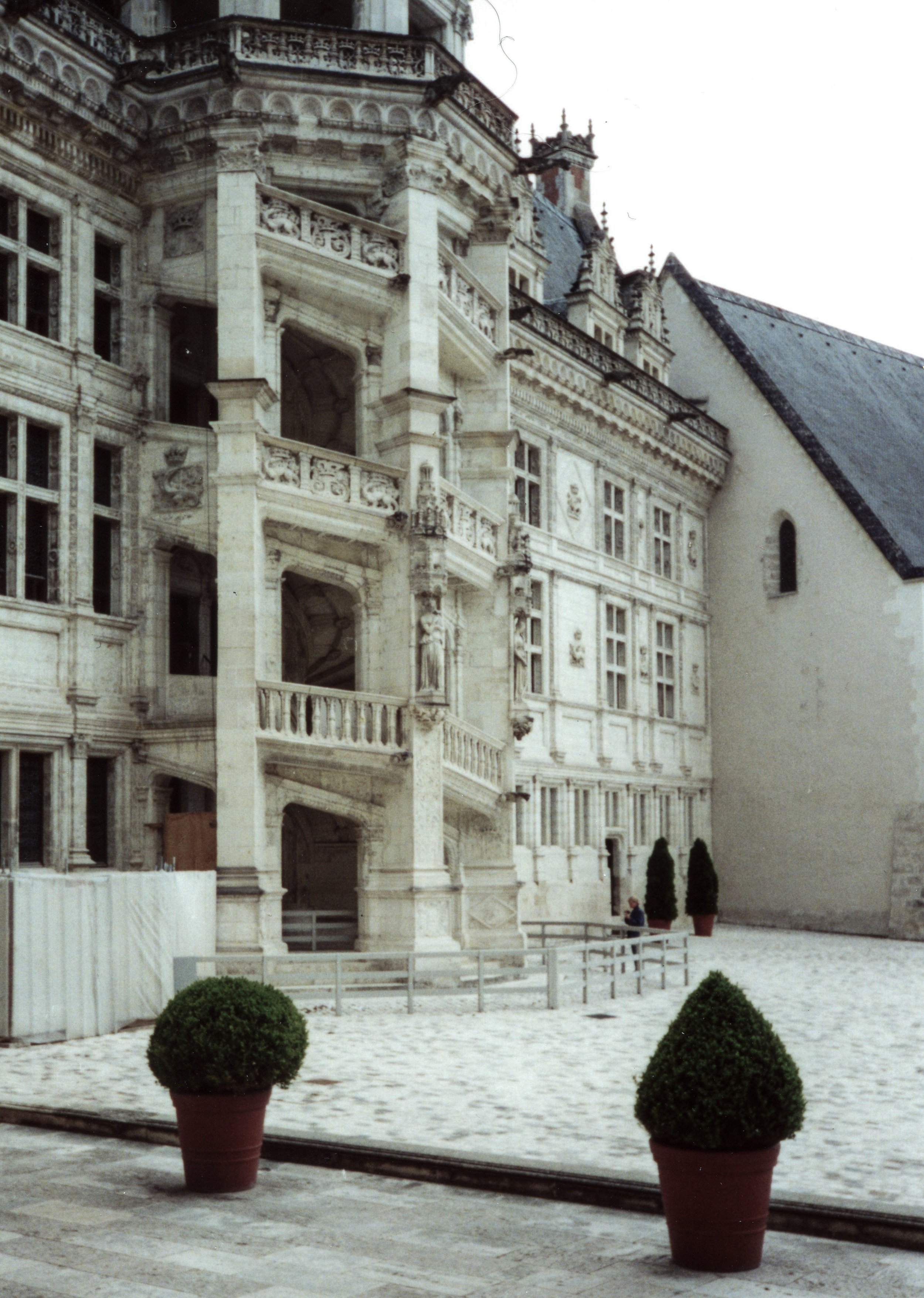 The photo shows an ornate, historic building with detailed stone architecture, several balconies, and large windows. In the foreground, there are potted plants and a cobblestone courtyard.