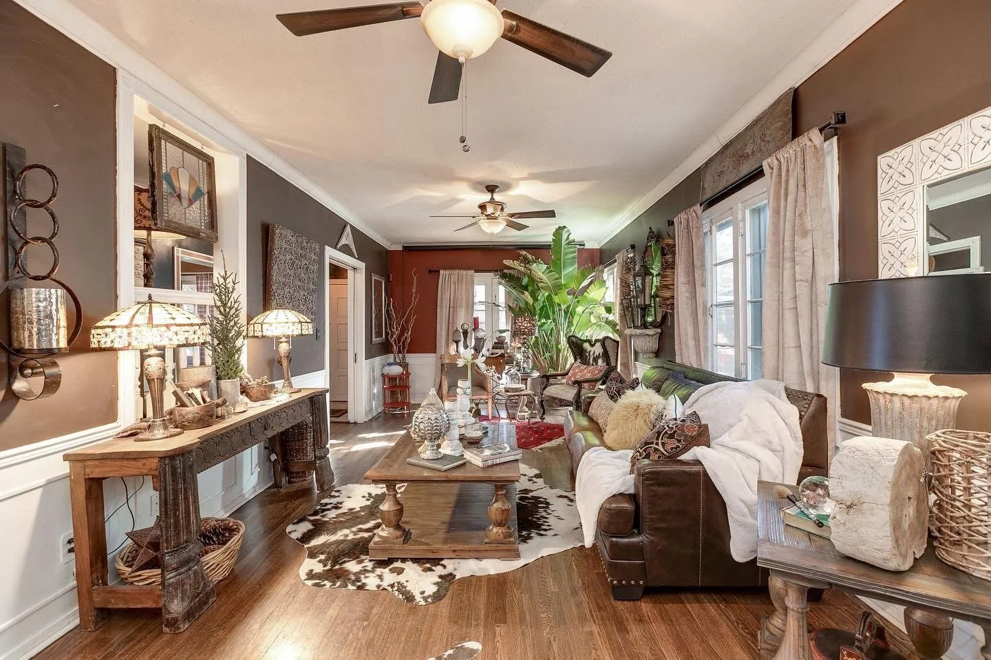 Living room with dark brown walls, white trim, hardwood floor, and multiple windows with beige curtains. Contains a dark leather sofa with pillows and a throw blanket, a wooden coffee table with decorative items, and a cowhide rug. There are large pl