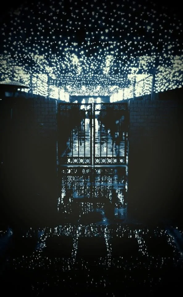 A dark outdoor scene with a lit canopy of small lights overhead and reflections on the wet ground, with a gate and brick walls in the background.