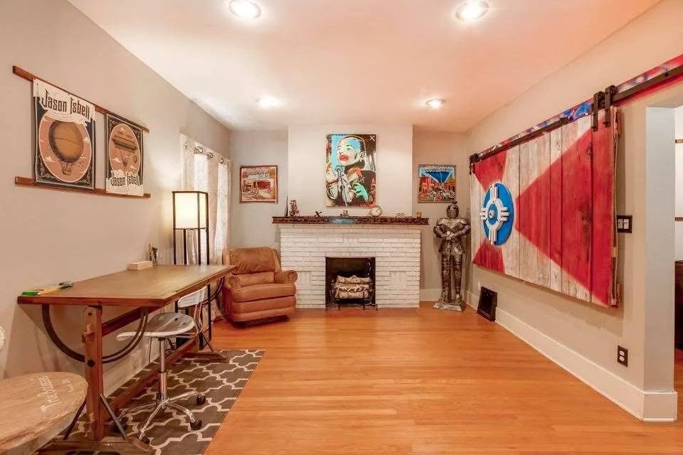 A living room with a white brick fireplace, artwork, and mounted decorative items. There is a brown armchair, a small desk with a chair, a colorful abstract painting, a statue of a knight, and a large red and beige geometric wall hanging with a blue 