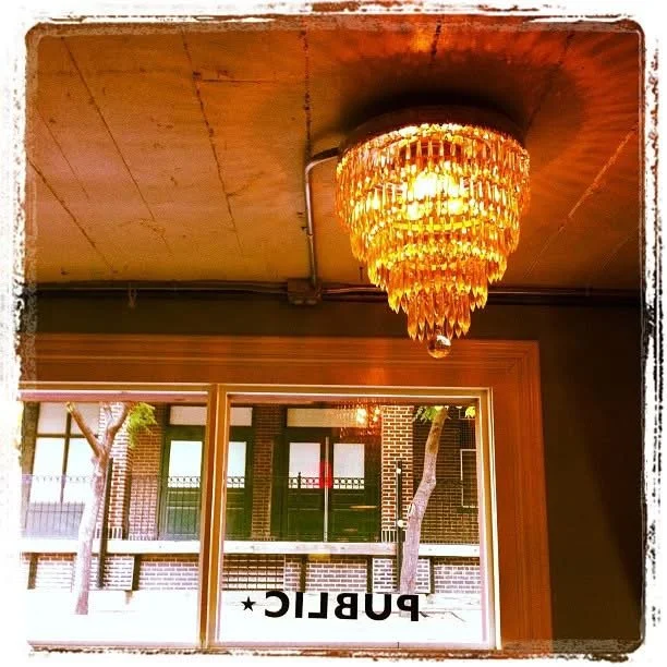 Interior view of a room with a large, crystal chandelier hanging from a wooden ceiling. A window in the background shows a brick building and trees outside, with the word 'PUBLIC' written upside down in the window.