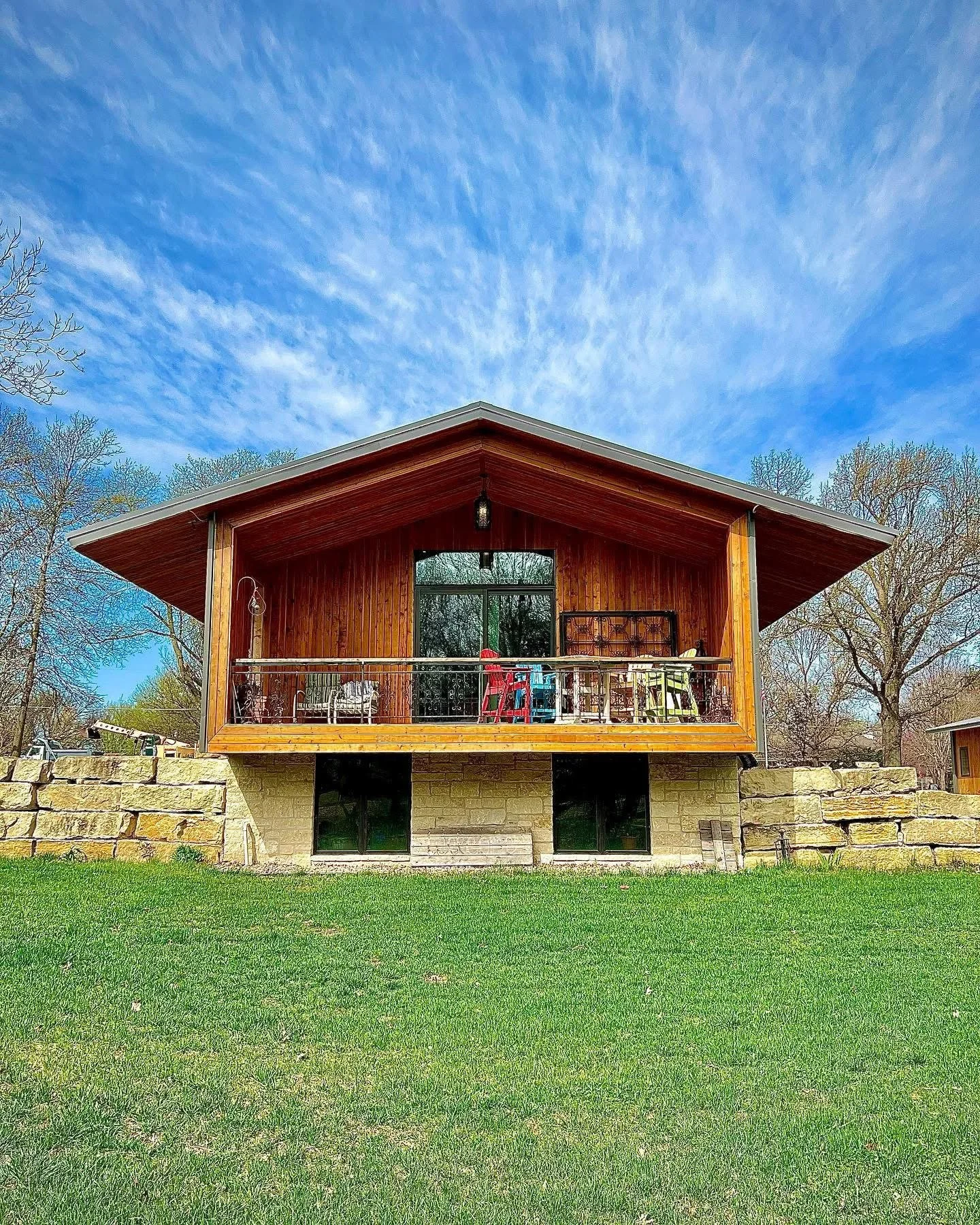 A two-story house with a wooden upper level and a stone lower level, surrounded by trees and a green lawn, under a blue sky with scattered clouds.