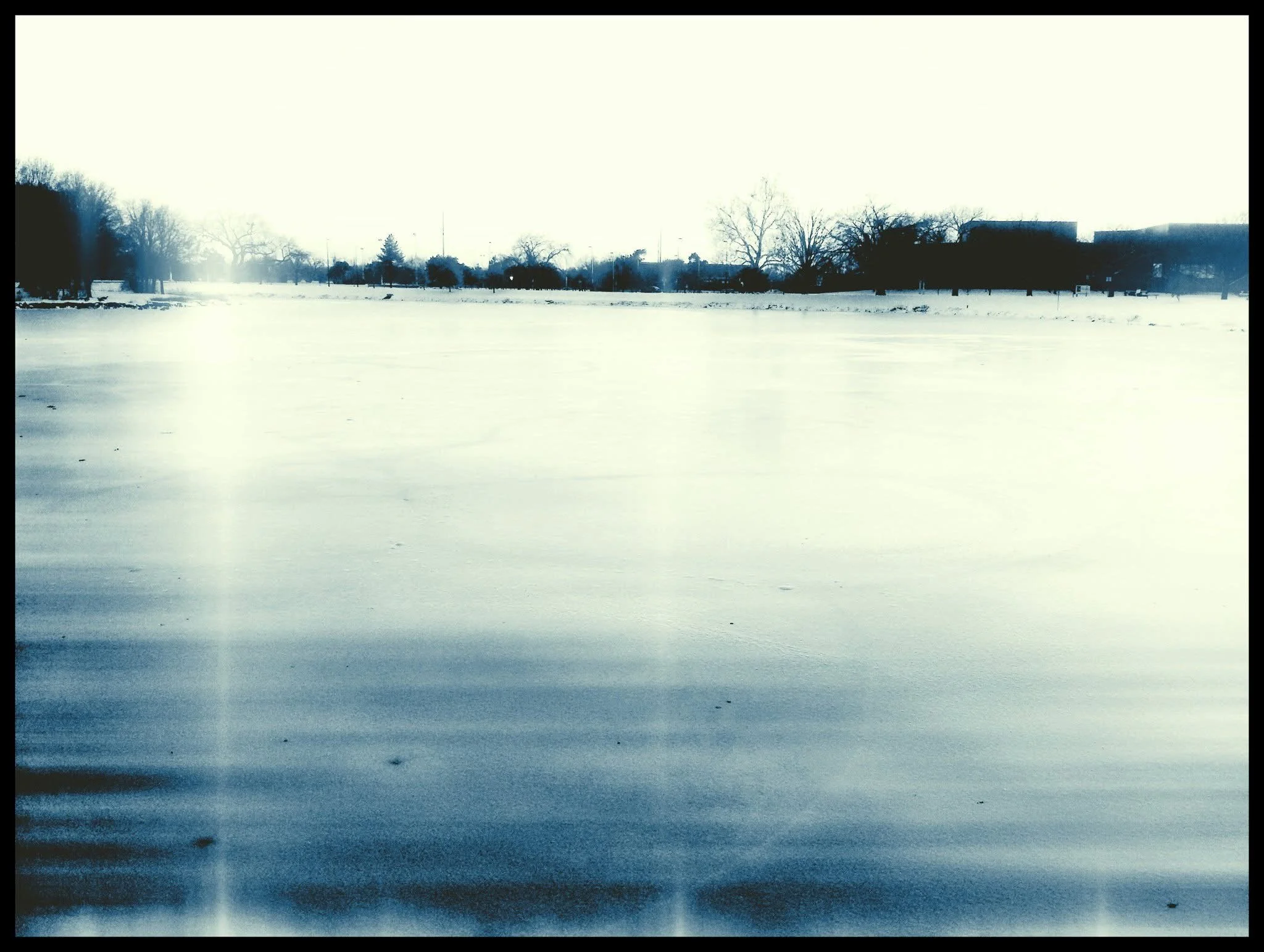 A snow-covered frozen lake with a tree-lined horizon and a few buildings in the distance, under a bright sky.