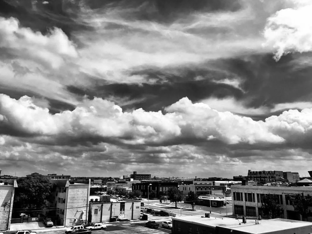 Black and white image of a cityscape with buildings and roads under a sky filled with large, dramatic clouds.
