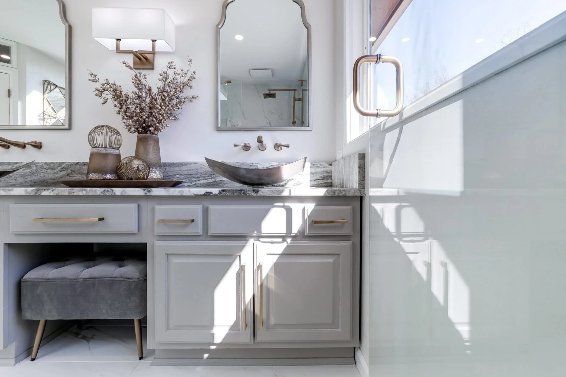 Modern bathroom vanity with a marble countertop, a vessel sink, and decorative vases with dried flowers in neutral tones. There is a mirror above the sink, and natural light is shining through a window on the right side.