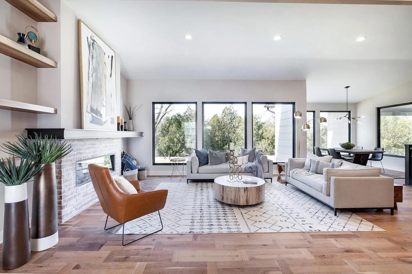 Bright and spacious living room with large windows, white sofas, a brown leather armchair, a round wooden coffee table, a patterned rug, and modern decor. View of greenery outside.