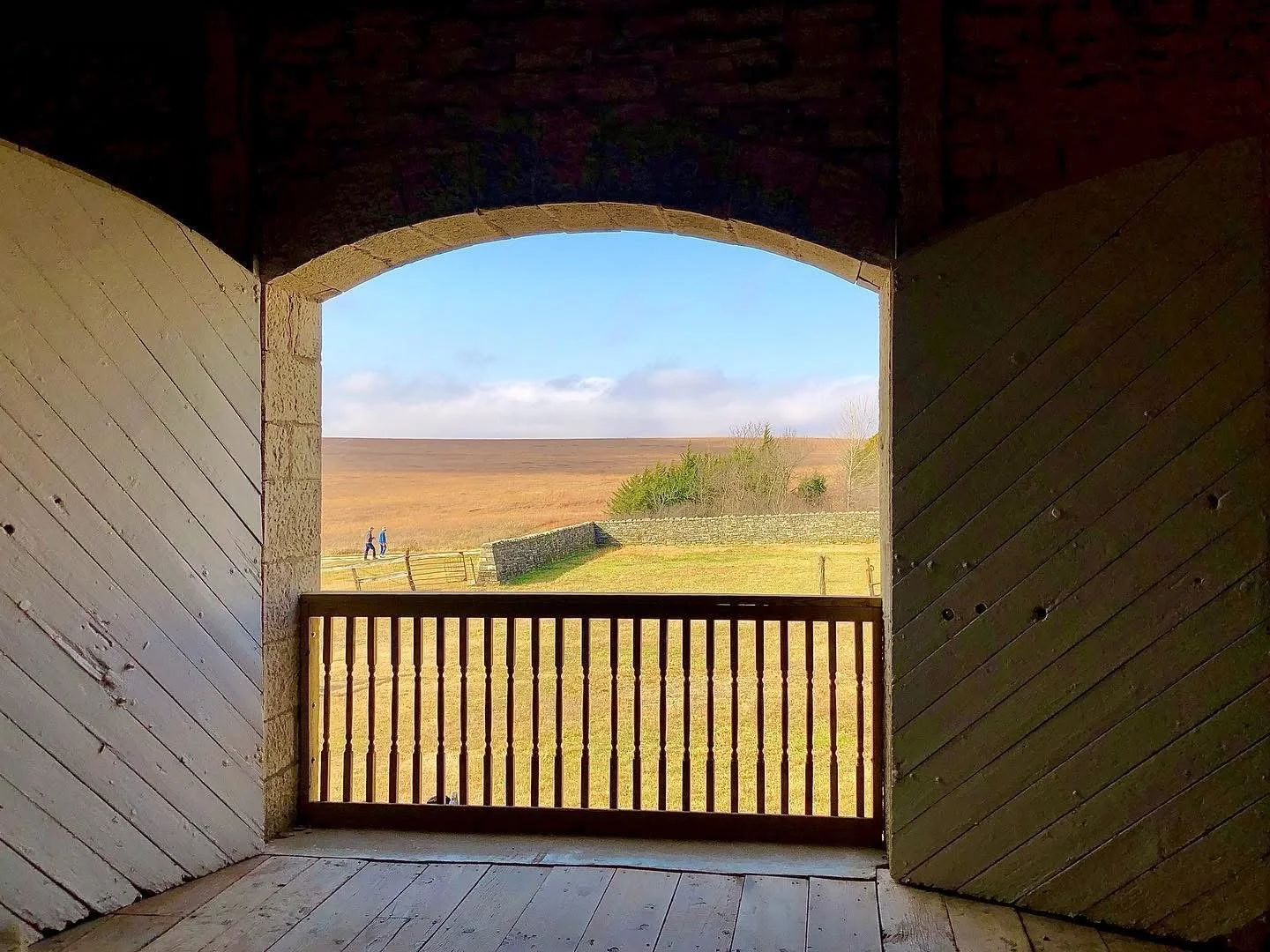 View through a wooden opening showing a grassy field, small stone wall, some trees, and a partly cloudy sky with two people walking in the distance.