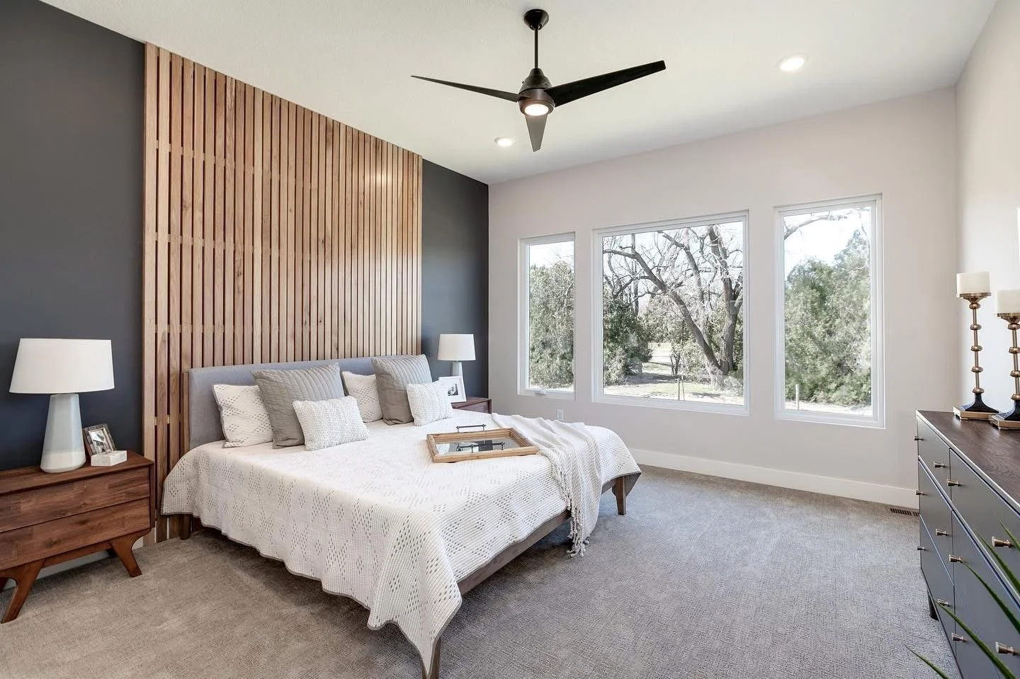 A modern bedroom with a large bed with white bedding, gray pillows, wooden nightstands with white lamps, a wooden headboard with vertical slats, large windows showing trees outside, and a ceiling fan.