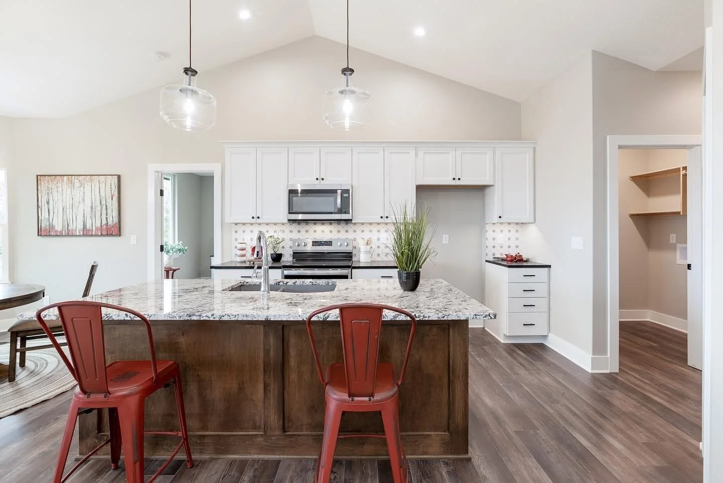 Modern kitchen with white upper cabinets, black countertops, stainless steel appliances, and a large island with a wooden base. Red metal chairs are placed around the island, and there are two pendant lights hanging from the ceiling. The room has a l