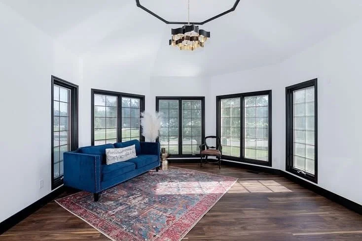 Bright room with white walls, multiple large windows, a blue sofa with pillows, an oriental rug, a single armchair, and a modern chandelier.