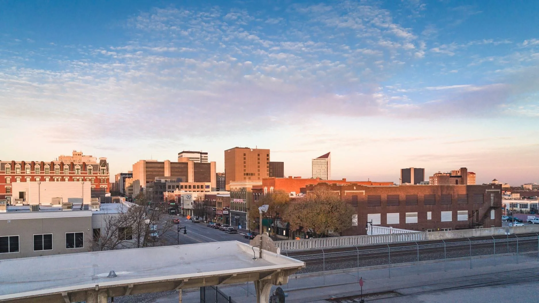 City skyline during sunrise with a mix of modern and historic buildings, a street with cars, trees, and train tracks in the foreground.