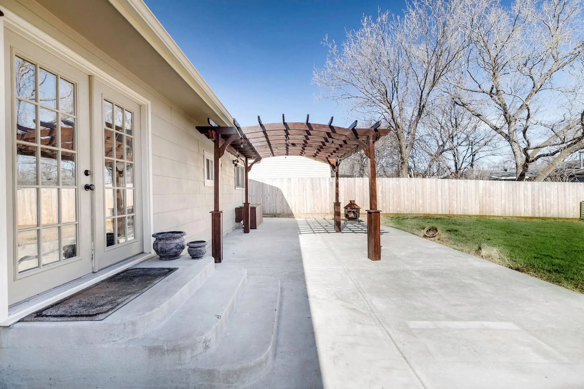 Back patio with a concrete floor, white house siding, glass double doors, potted plants, wooden pergola, outdoor fireplace, grassy yard, and large trees with bare branches.