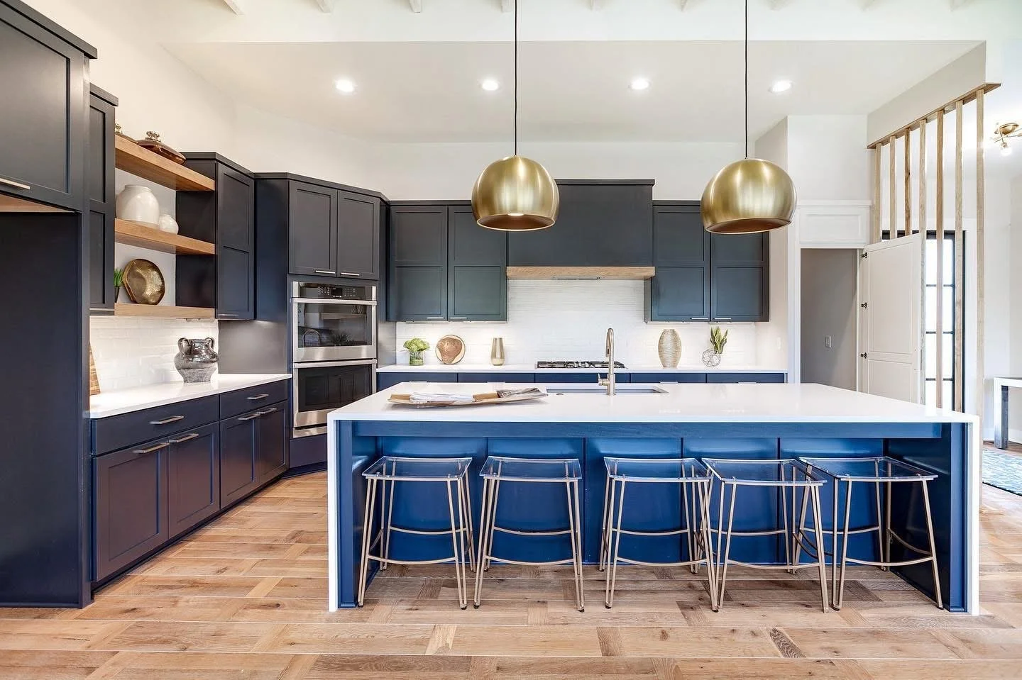 Modern kitchen with navy blue cabinets, white island, and gold pendant lights, wooden flooring, decorated with vases and decorative items.