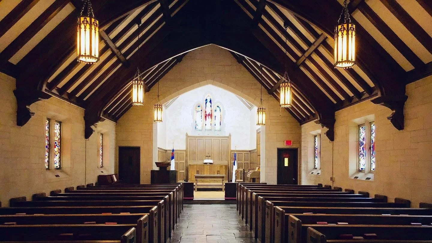 Interior of a church with wooden pews, stained glass windows, hanging lantern lights, and an altar with religious symbols at the front.