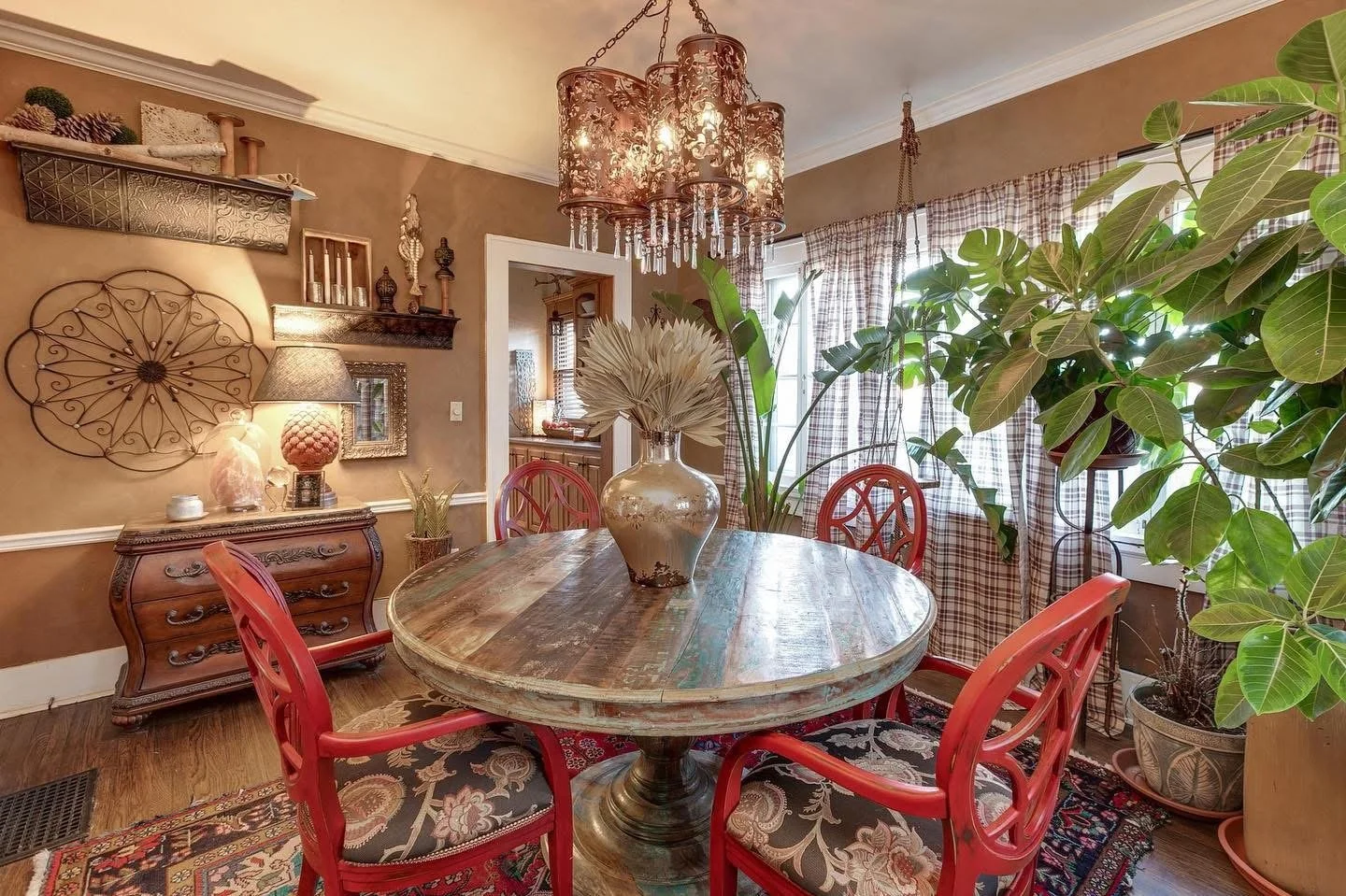 A dining room with a round wooden table, red chairs, large green plants, a chandelier, and vintage decor on the walls.
