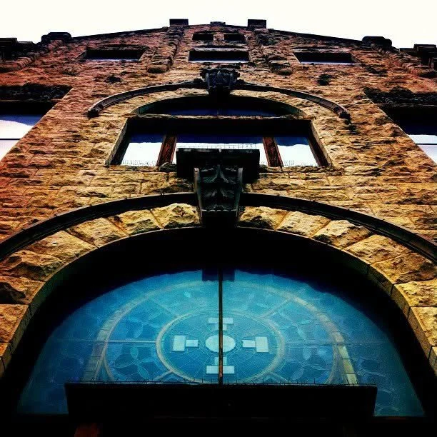 Close-up view of a historic brick building with arched windows and decorative stone details, featuring a clock with a blue face and Roman numerals.