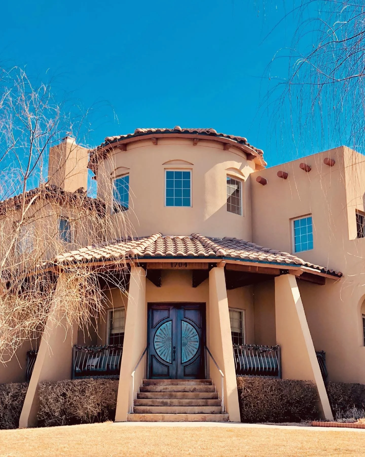 A large beige stucco house with a red-tile roof, multiple windows, and a large front door with stairs leading up to it, set against a clear blue sky and some leafless trees.