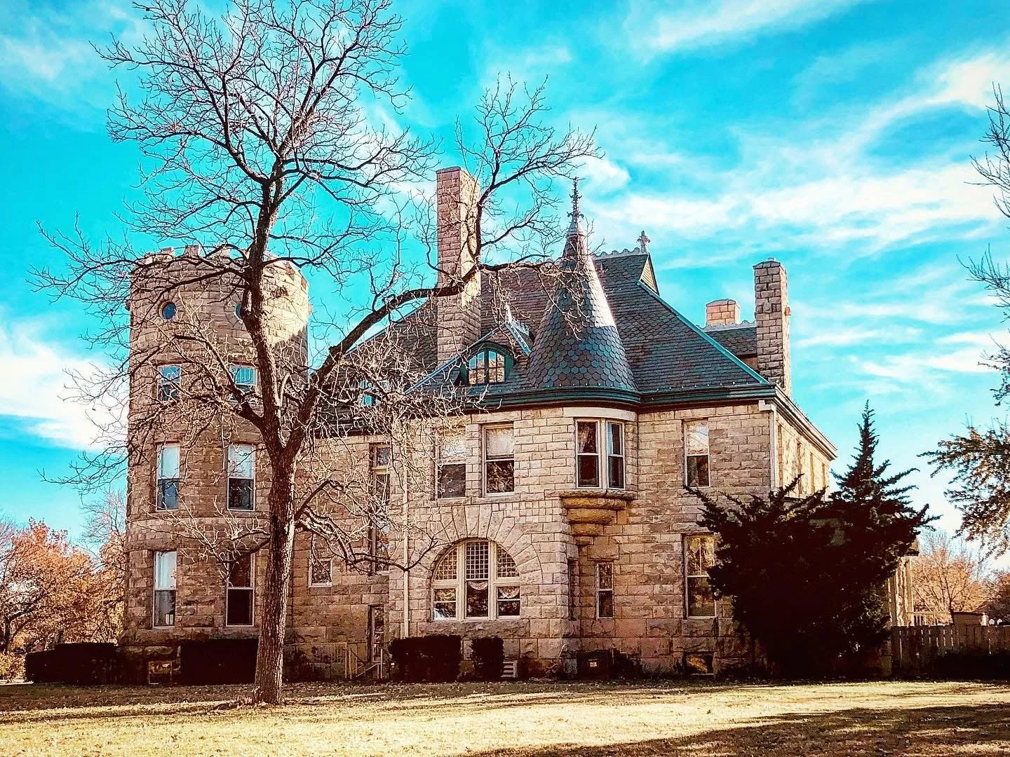A historic stone mansion with turrets, pointed roofs, and multiple chimneys surrounded by trees and a grassy lawn under a blue sky.