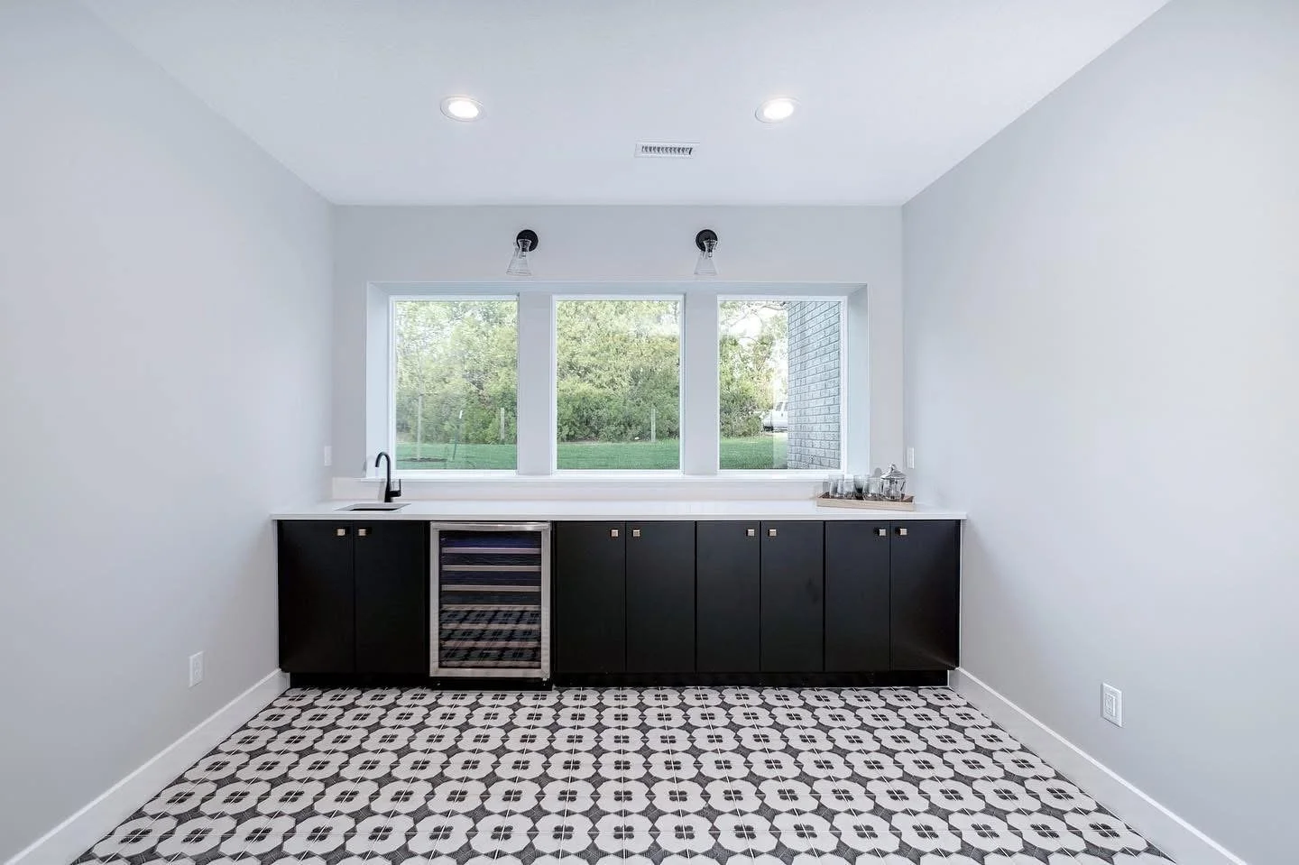 Modern kitchen with black cabinetry, a built-in wine cooler, white countertops, and a large window overlooking a green outdoor space, with black and white patterned tile floor.
