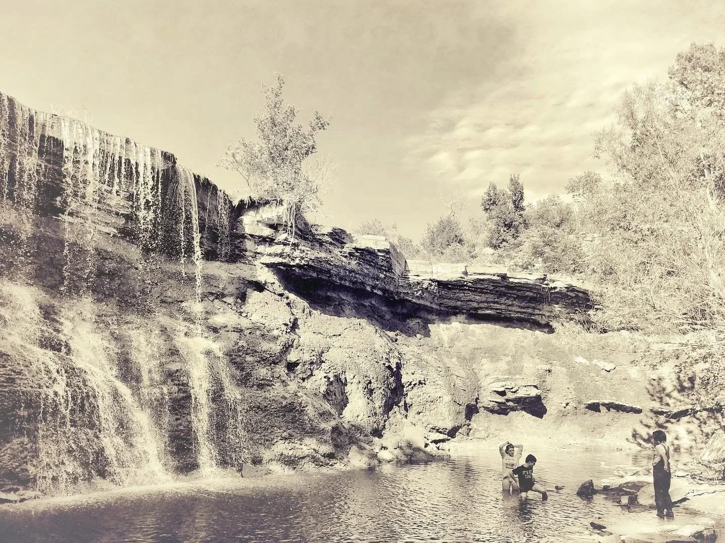 Children playing in a small natural waterfall and pool surrounded by rocky cliffs and trees, with a cloudy sky overhead.