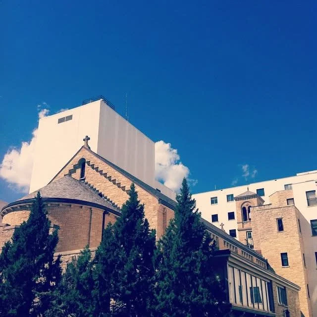 A church with a brick roof and cross on top, surrounded by modern buildings and green trees under a blue sky with some clouds.