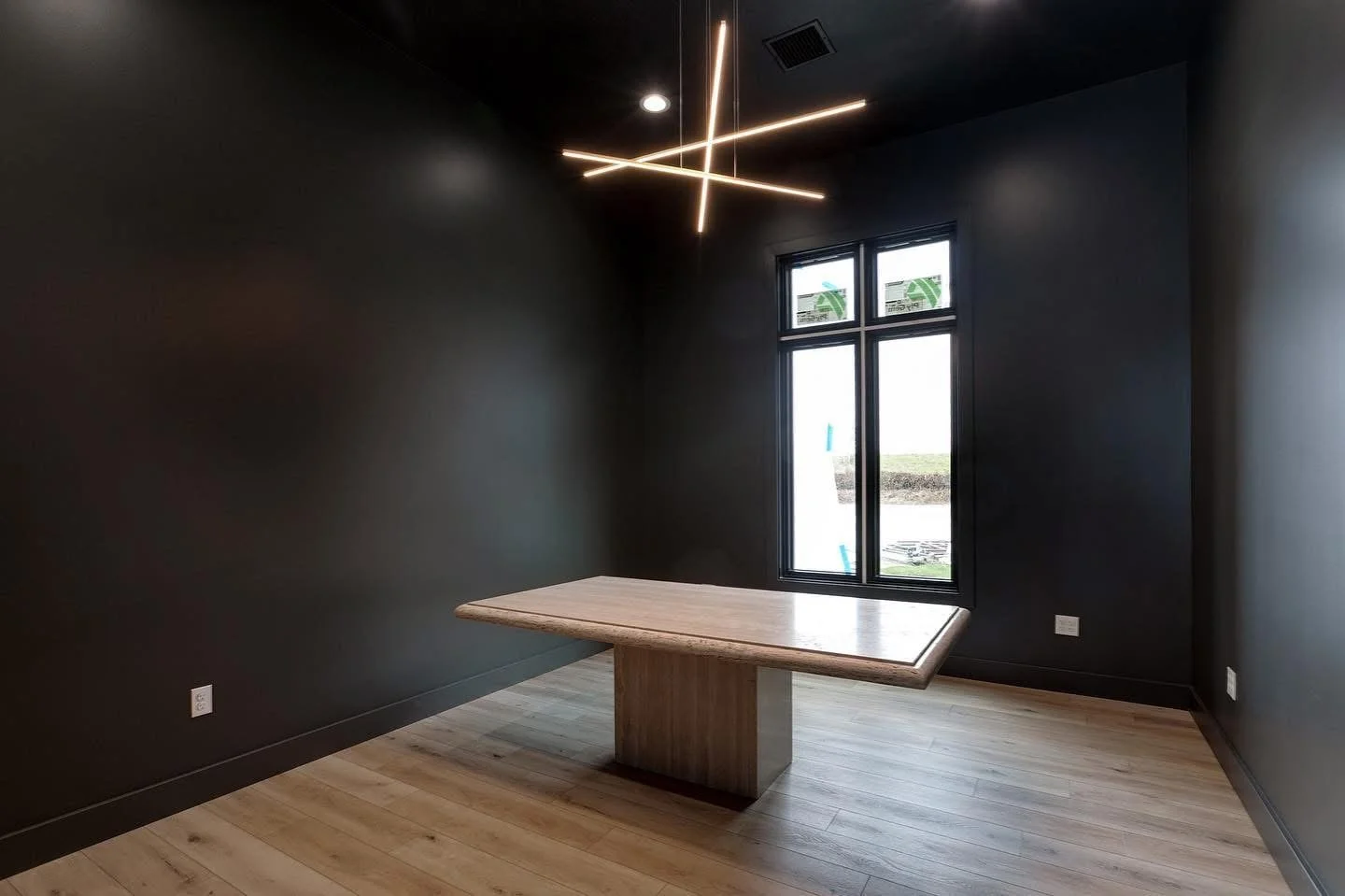 Empty room with dark walls, light wooden floor, a single wooden table, a window, and modern ceiling light fixture.