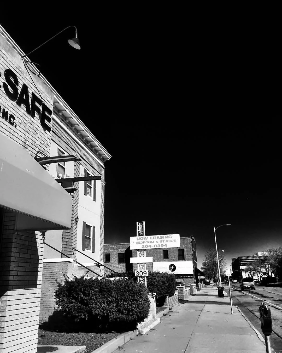 Black and white photo of a sidewalk in front of buildings, with a sign advertising leasing for a studio apartment, and a portion of a building that has a large sign with the word 'SAFE' visible.