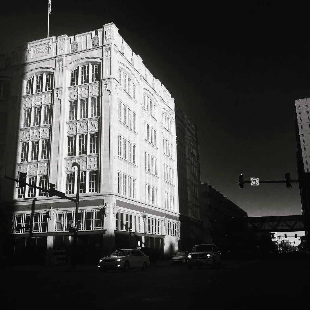 Black and white photo of a tall, ornate building with multiple windows, streetlights, cars on the street, and traffic signals, with a shadow cast across it.