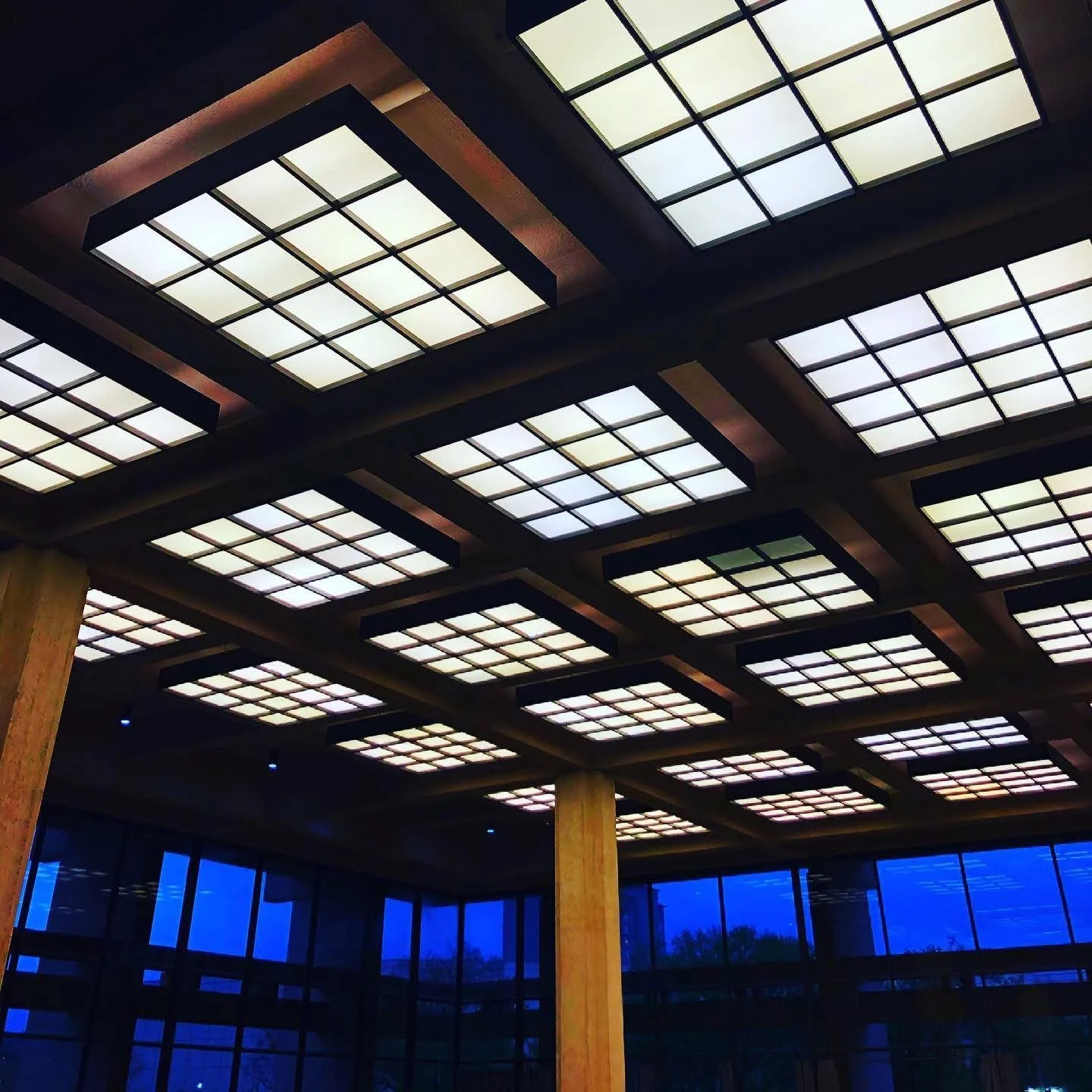 Ceiling with illuminated square panels, wooden support beams, and a large blue-tinted window in the background.