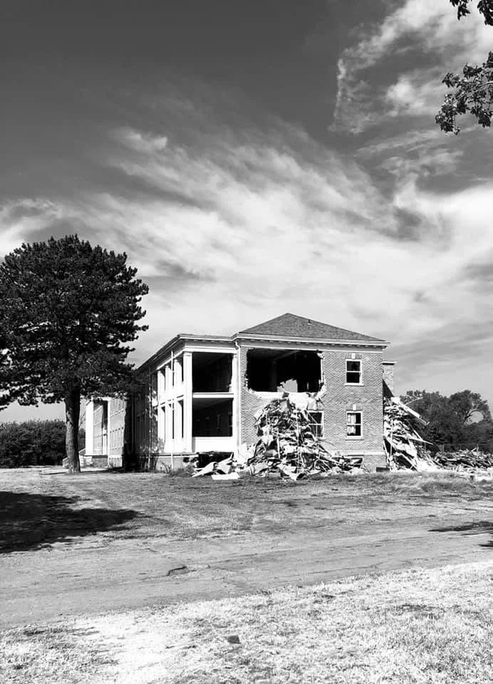 A black and white photo of a two-story house under construction or renovation, with part of the front facade demolished, exposing the interior. There is a large tree on the left side and a cloudy sky overhead.