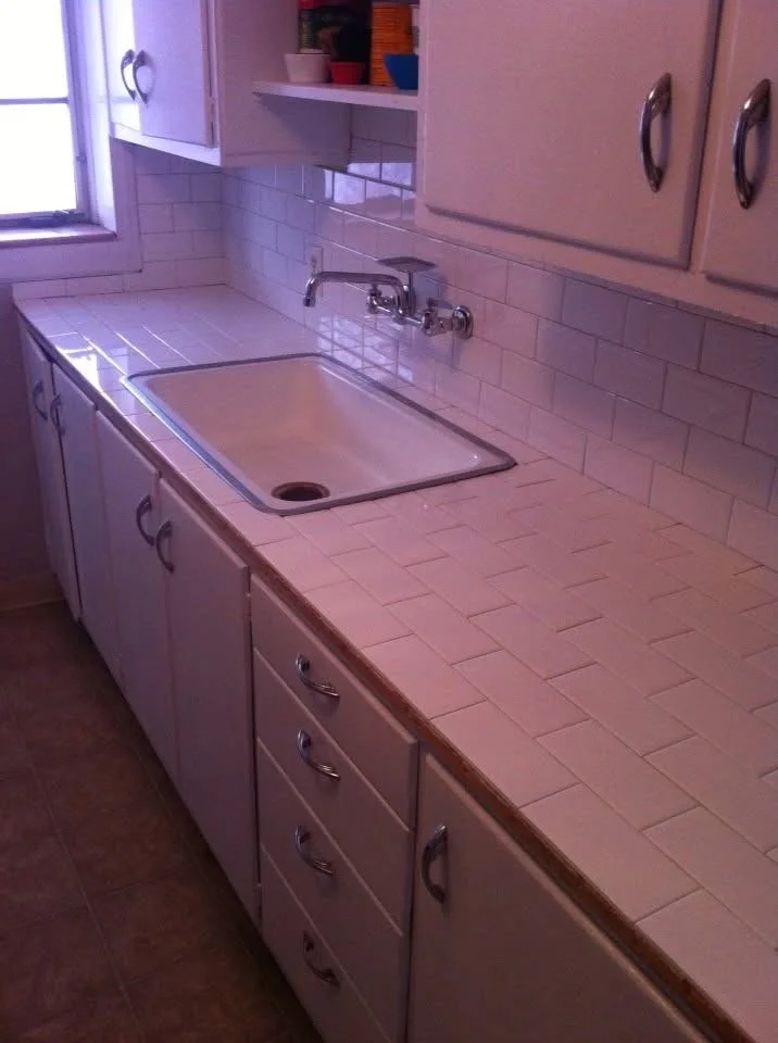 Kitchen sink area with a white tiled countertop, upper and lower cabinets, and a window on the left side.
