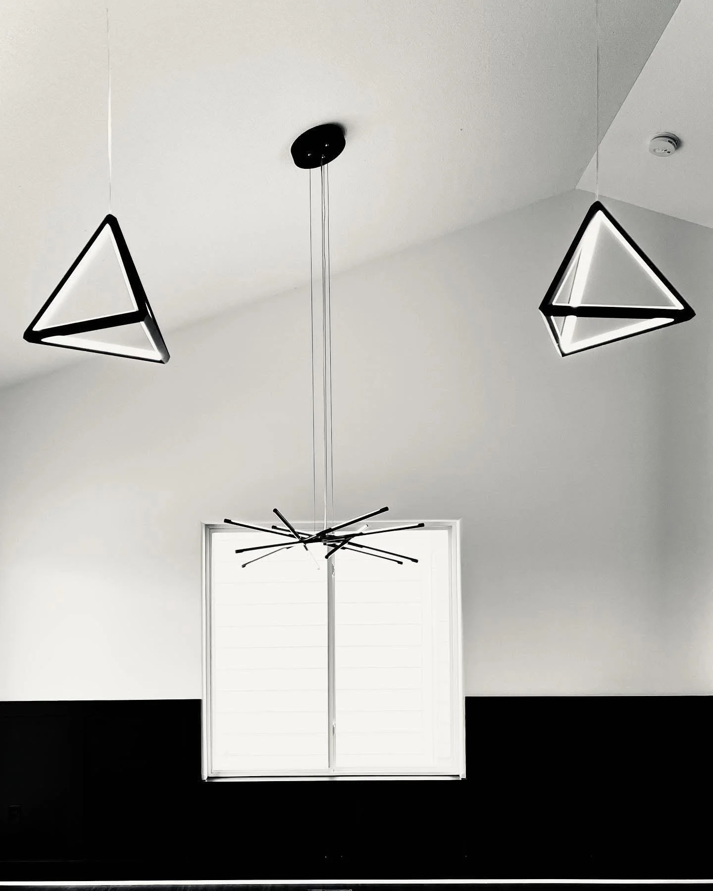 Modern interior with two geometric pendant lights hanging from the ceiling and a contemporary chandelier below a window with closed blinds.