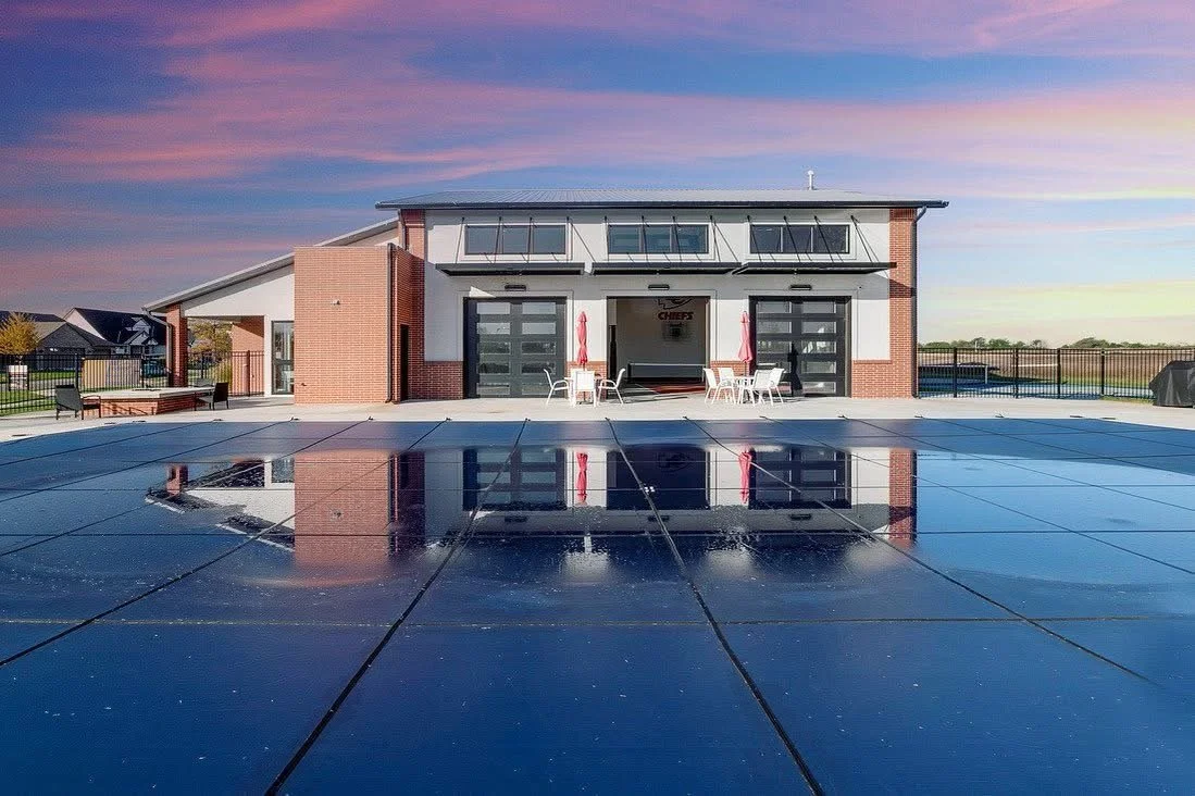 A modern building with a garage, outdoor seating, and a large reflecting pool on a sunny day with pink and purple sky.