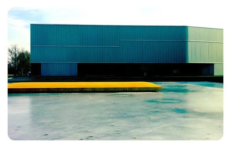Modern building with a gradient blue and green exterior, parking lot in foreground, and a yellow and black speed bump in the parking area.
