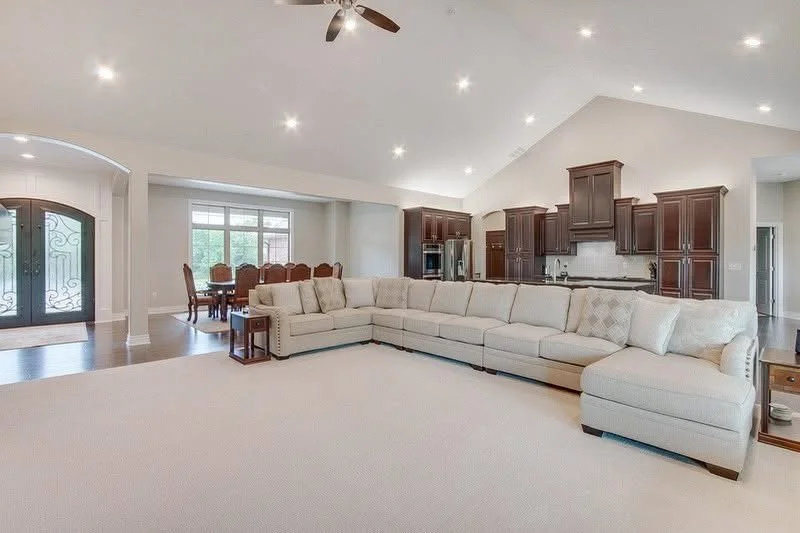 Spacious living room with white sectional sofa, wooden dining table, and kitchen with dark wooden cabinets.