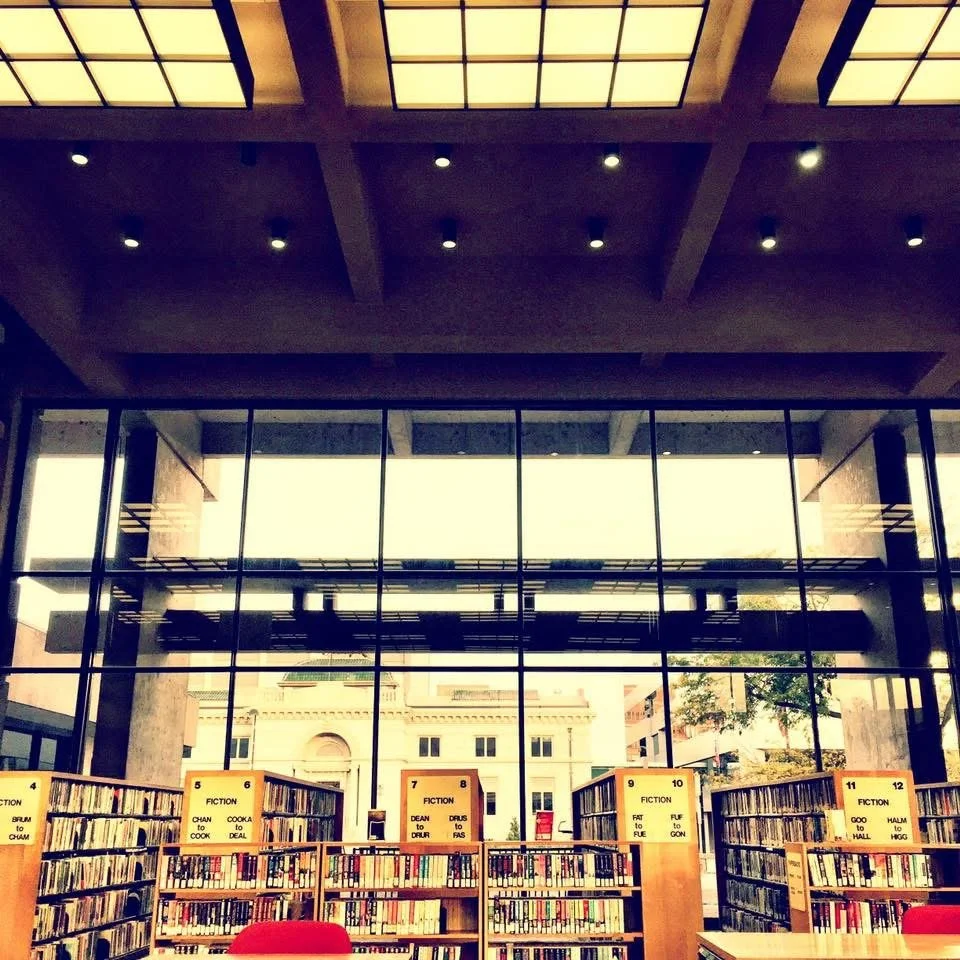 Interior of a modern library with large glass windows, wooden bookshelves filled with books, and labels indicating fiction sections numbered 4 to 12.