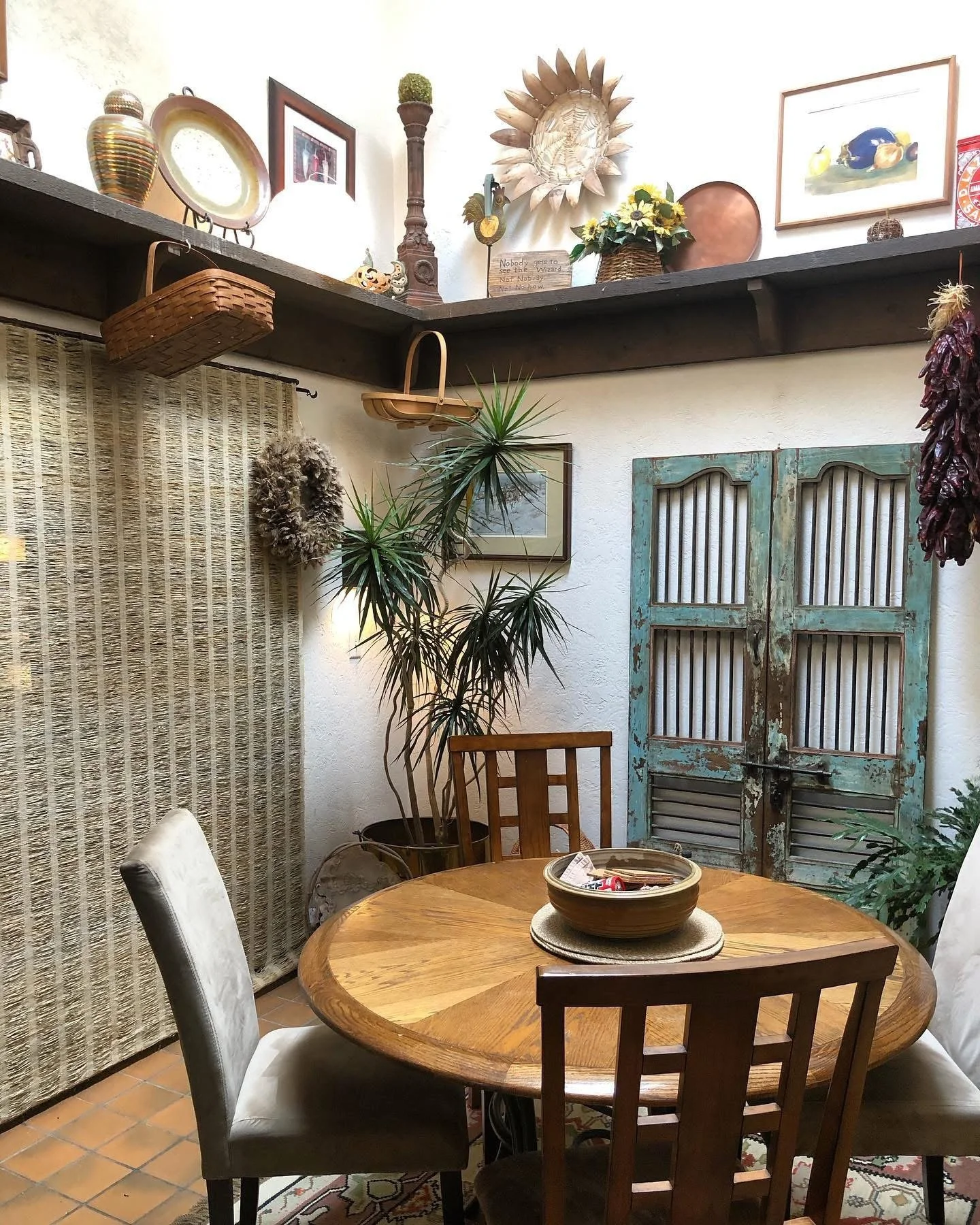 Cozy dining nook with a circular wooden table surrounded by mixed chairs, large potted plant, vintage distressed double doors, decorative wall art, shelves with baskets, and eclectic decorative items on upper shelves.