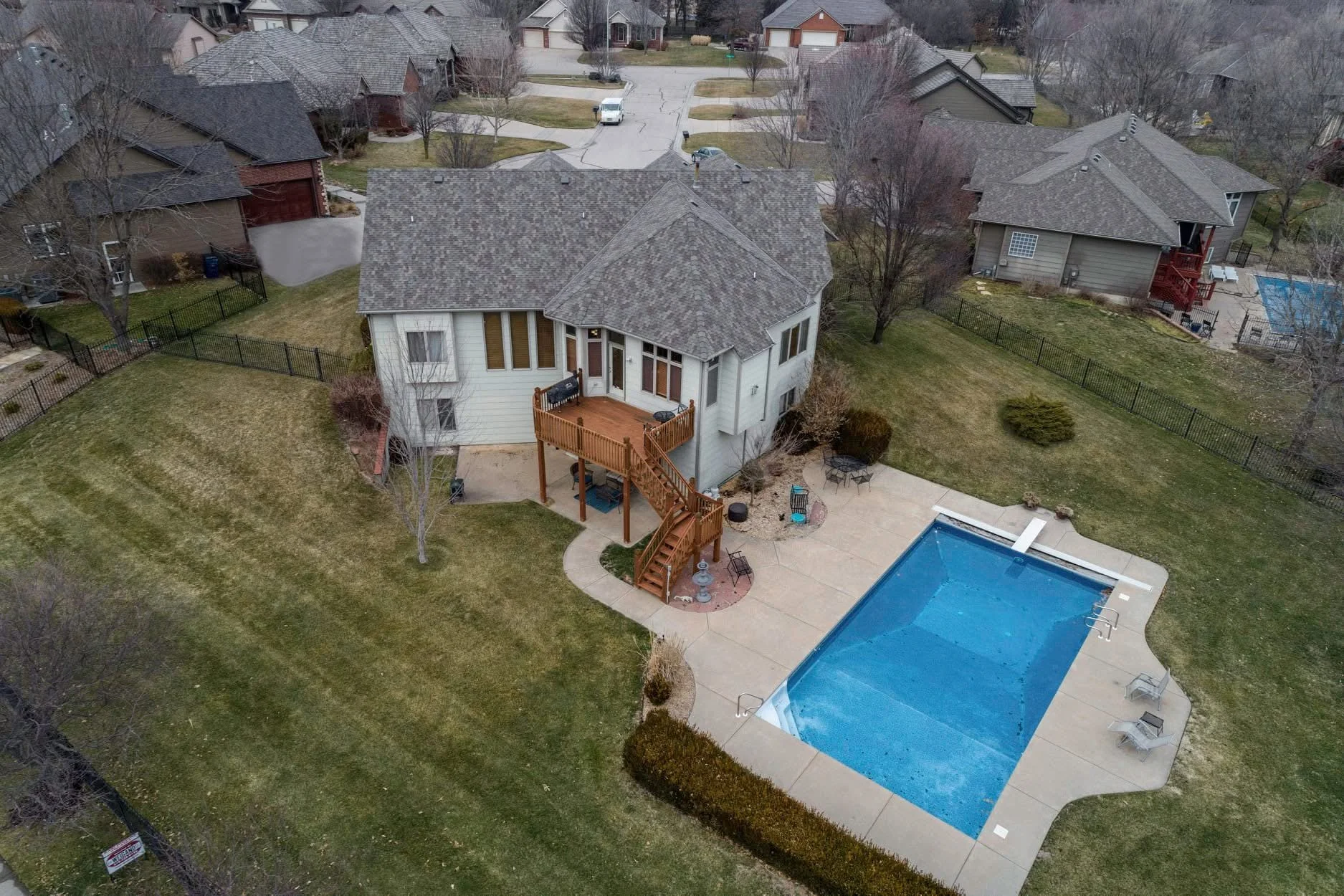An aerial view of a suburban backyard featuring a large in-ground swimming pool with a cover partially rolled back, surrounding concrete patio with outdoor furniture, a wooden deck attached to a white two-story house, and a well-kept lawn with trees 