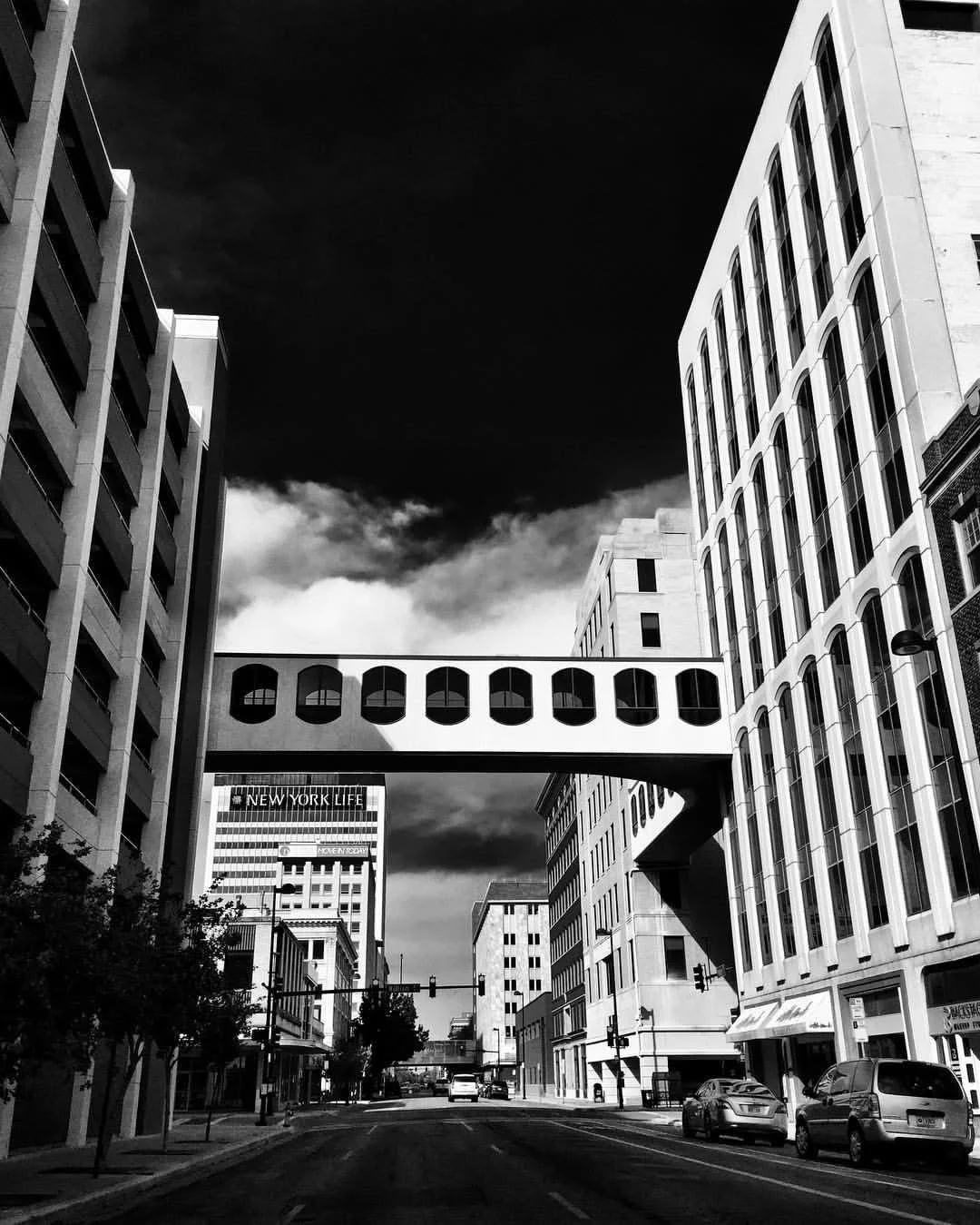 Black and white photo of a city street with tall buildings, a pedestrian overpass, and a cloudy sky