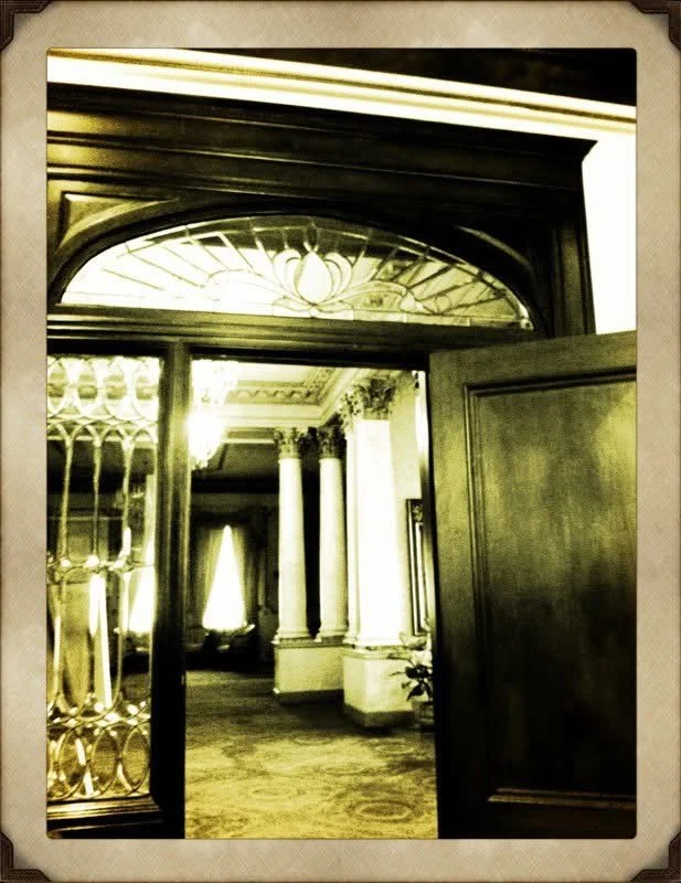 Open wooden door leading into an elegant, historic interior with tall white columns, ornate ceiling, and chandeliers.
