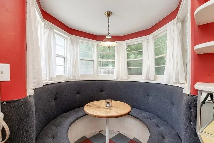 A cozy bay window nook with white curtains, a round wooden table, and a black cushioned bench seating. A vintage hanging light fixture is centered above the table, and there are white shelves on the right side. The walls are painted red.